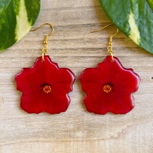 Puede incluir: Un par de pendientes de resina roja con forma de flores de hibisco y ganchos dorados.