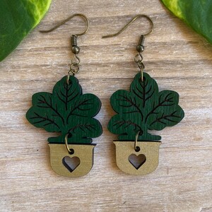 Puede incluir: Pendientes de madera con un diseño de planta verde en una maceta dorada con un recorte en forma de corazón. Los pendientes tienen ganchos para las orejas de color bronce y están sobre un fondo de madera.