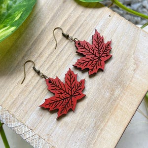 May include: A pair of red maple leaf earrings with bronze hooks. The leaves are made of wood and have a detailed engraved design.