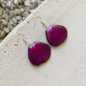 May include: A pair of dangle earrings with a deep purple resin petal shape. The earrings have silver hooks.