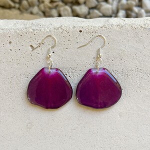 May include: A pair of earrings featuring a purple flower petal design. The earrings are made of resin and have silver hooks.