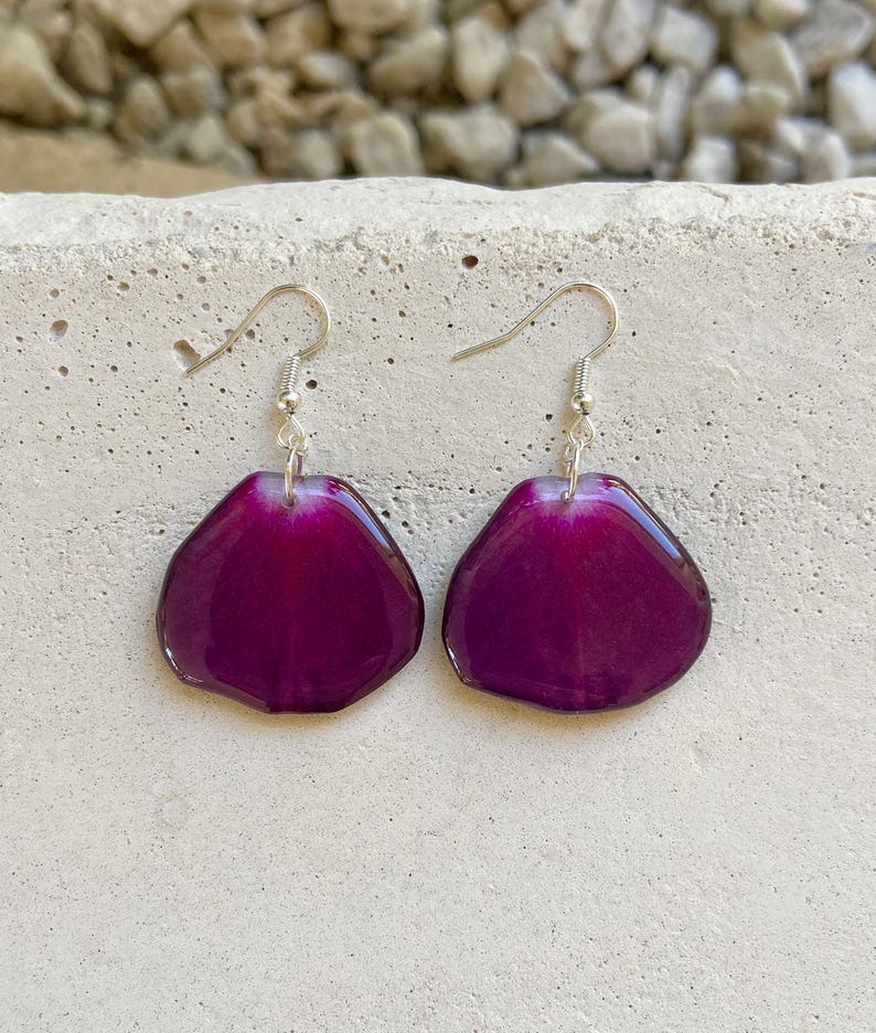 May include: A pair of earrings featuring a purple flower petal design. The earrings are made of resin and have silver hooks.
