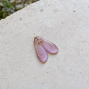 Puede incluir: Un delicado colgante con un par de semillas de arce, hecho de resina y pintado en un suave tono morado.