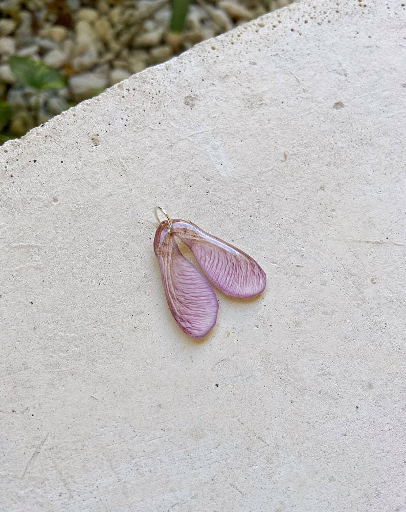 Puede incluir: Un delicado colgante con un par de semillas de arce, hecho de resina y pintado en un suave tono morado.
