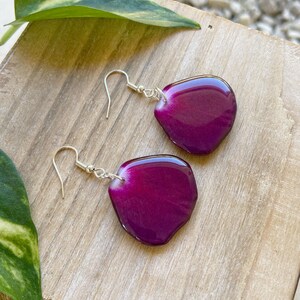 May include: A pair of earrings featuring a purple resin petal design. The earrings are made with silver hooks.