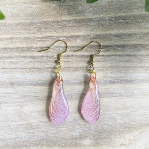 May include: A pair of earrings featuring delicate pink maple seed wings with gold hooks.