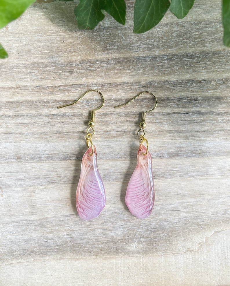 May include: A pair of earrings featuring delicate pink maple seed wings with gold hooks.