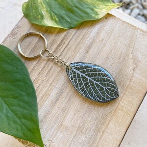 May include: A silver key chain with a dark green leaf-shaped resin charm. The leaf has a white vein pattern.