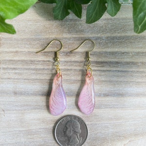 May include: A pair of delicate earrings featuring pink and white resin maple seed wings with gold hooks.