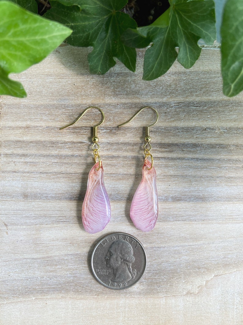 May include: A pair of delicate earrings featuring pink and white resin maple seed wings with gold hooks.