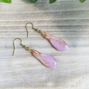 May include: A pair of earrings featuring delicate pink resin maple seed wings with gold-toned hooks.