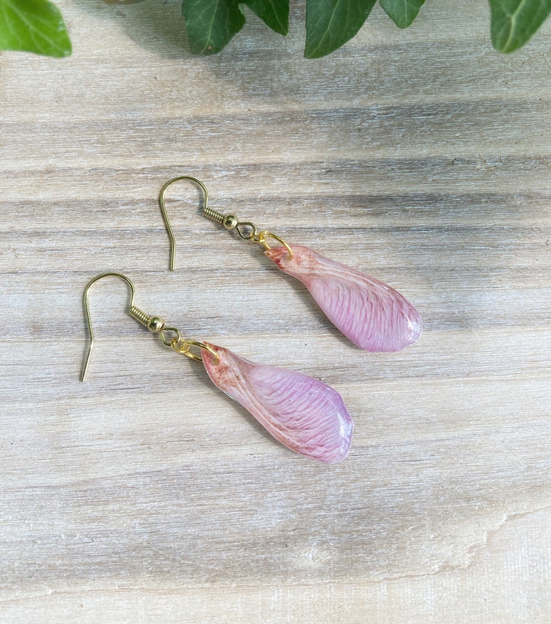 May include: A pair of earrings featuring delicate pink resin maple seed wings with gold-toned hooks.