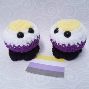 May include: Two handmade plush toys with a non-binary pride flag. The toys are round with yellow, white, purple, and black colors. The toys have large black eyes. A small non-binary flag is placed in front of the toys.