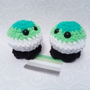 May include: Two crocheted plushies shaped like blobs with black eyes and a green, white, and black color scheme, reminiscent of the asexual pride flag. The plushies are sitting on a white surface with a small asexual pride flag card in front of them.