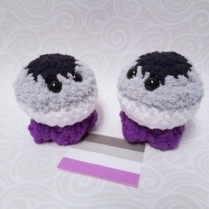 May include: Two gray, white, and purple crocheted plushies with black eyes. The plushies are sitting on a white surface with an asexual pride flag behind them.