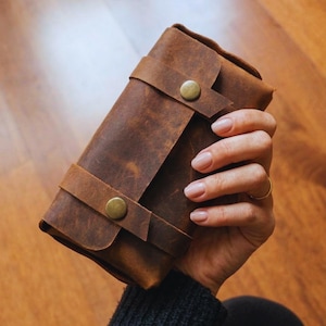 May include: A brown leather wallet with a distressed finish. The wallet is secured with two leather straps and brass-coloured button closures. The leather has a textured appearance, and the wallet is held in a hand.
