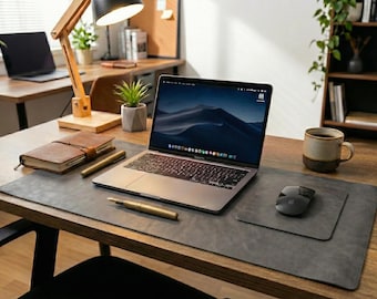 May include: A dark gray leather desk mat featuring a rectangular shape, placed on a wooden desk. The mat serves as a base for a silver laptop, a black wireless mouse, a gold fountain pen, a brown leather notebook, and a ceramic coffee mug.
