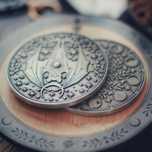 May include: Two silver-colored, detailed coins featuring celestial and bat designs. One coin displays a bat with outstretched wings, surrounded by stars. The other coin shows phases of the moon and floral elements. The coins rest on a wooden surface.