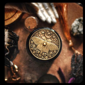 Puede incluir: Una caja redonda negra y dorada con un dise&ntilde;o detallado de una polilla, una luna creciente y estrellas. La caja es de metal y tiene una est&eacute;tica vintage.