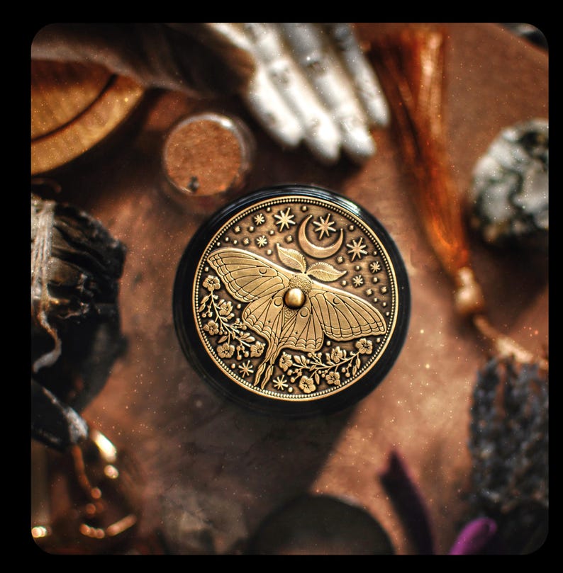 Puede incluir: Una caja redonda negra y dorada con un dise&ntilde;o detallado de una polilla, una luna creciente y estrellas. La caja es de metal y tiene una est&eacute;tica vintage.