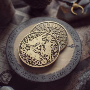 以下が含まれることがあります： 金色の丸いコイン2枚。ウサギと花の模様が細かく彫刻されています。コインは、暗い灰色の縁が付いた装飾的な円形トレイ内の木製の表面に置かれています。全体的な美学はヴィンテージで芸術的です。