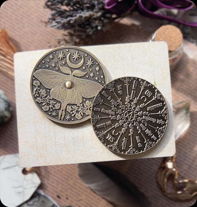 Puede incluir: Dos amuletos de metal dorado antiguo. Un amuleto presenta una ilustraci&oacute;n detallada de una polilla con una luna creciente y estrellas. El otro amuleto es una rueda circular con las palabras "dejar ir", "el tiempo es correcto", "nueva direcci&oacute;n", "no te preocupes", "s&iacute;", "improbable", "considerar", "reconciliarse", "ir ahora", "no es correcto", "direcci&oacute;n correcta", "continuar", "y "todav&iacute;a no" escritas alrededor del borde.