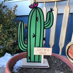 May include: A green and black cactus-shaped wooden yard decoration with a pink flower on top. The sign reads "DON'T BE A PRICK". The decoration is in a terracotta pot with soil.