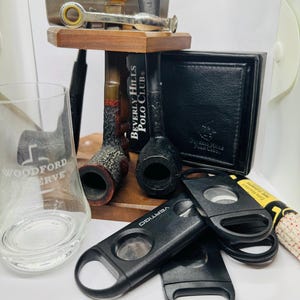 May include: An assortment of smoking accessories, including two tobacco pipes, a black leather wallet, and two black cigar cutters. A glass with "Woodford Reserve" is visible, along with a metal and wood pipe stand.