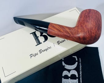 May include: A brown and black pipe with a curved design, resting on a box and a black pouch. The box reads "Pipe Bruyère" and "Since 1858". The pouch has the letters "BC" printed on it.