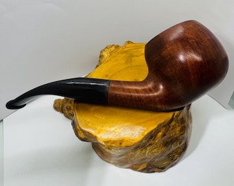 May include: A brown wooden pipe with a curved black stem. The pipe rests on a yellow and brown wooden base. The pipe has a smooth, polished finish and a rounded bowl.
