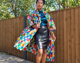 Op de afbeelding: Een lange, aan de voorzijde open kimono-jas met een levendig, veelkleurig geometrisch patchworkpatroon. Het wordt gedragen over een mouwloze top van donkergroenblauw satijn en een zwarte kokerrok van kunstleer met een split aan de zijkant.