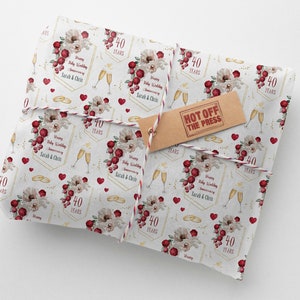 May include: White wrapping paper with a repeating pattern of gold rings, red hearts, and floral bouquets. The paper features the text "Happy Ruby Wedding Anniversary Sarah & Chris" and "40 Years" in a gold frame. The text "Hot Off The Press" is printed on a brown tag.