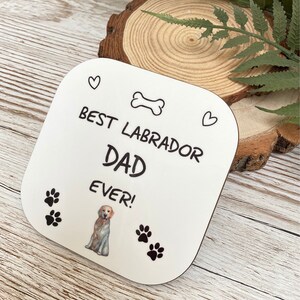 Może przedstawiać: Biały kwadratowy podkładka z czarnym tekstem, kt&oacute;ry brzmi "Best Labrador Dad Ever!" z czarną i białą kością, dwoma sercami i odciskami łap. Kresk&oacute;wkowy labrador retriever znajduje się w środku podkładki.