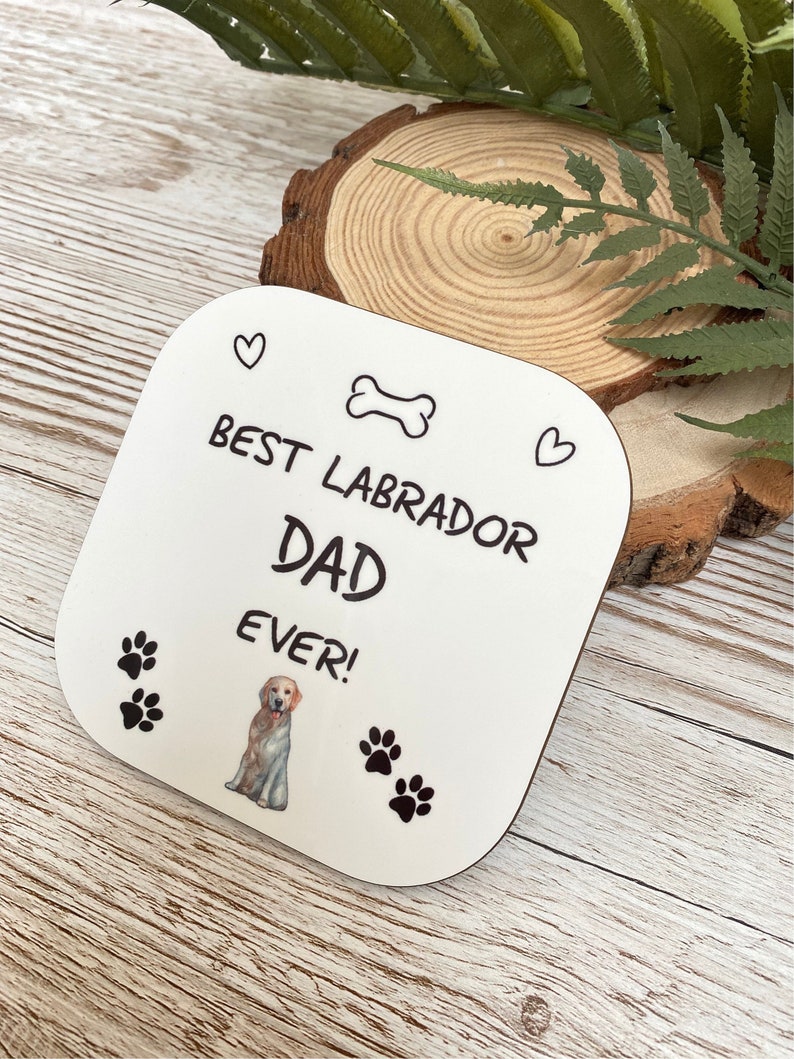 Może przedstawiać: Biały kwadratowy podkładka z czarnym tekstem, kt&oacute;ry brzmi "Best Labrador Dad Ever!" z czarną i białą kością, dwoma sercami i odciskami łap. Kresk&oacute;wkowy labrador retriever znajduje się w środku podkładki.
