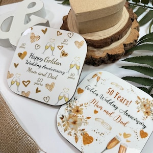 two coasters with gold and white designs celebrating a golden wedding anniversary. The coasters are placed on a wooden surface, surrounded by a burlap cloth and some greenery.