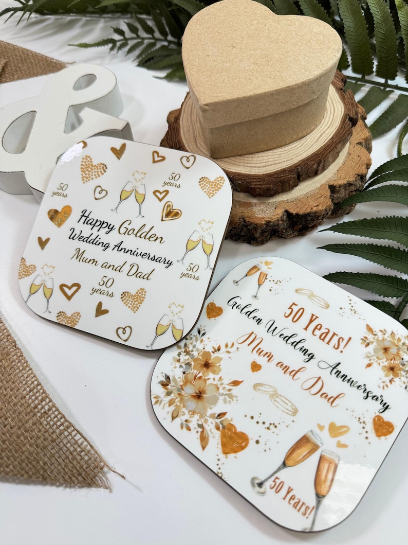 two coasters with gold and white designs celebrating a golden wedding anniversary. The coasters are placed on a wooden surface, surrounded by a burlap cloth and some greenery.