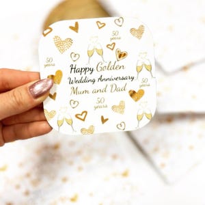 A hand holding a white card with gold designs and text, celebrating a golden wedding anniversary.