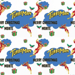 Puede incluir: Papel de regalo blanco con un patr&oacute;n repetido con el texto "TURBOMAN" en amarillo, un superh&eacute;roe rojo y amarillo, cajas de regalo verdes y las frases "MERRY CHRISTMAS" y "MONTE" en negro.