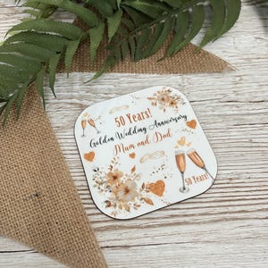 A 50th anniversary coaster with a floral design and champagne glasses, placed on a wooden surface with green leaves in the background.