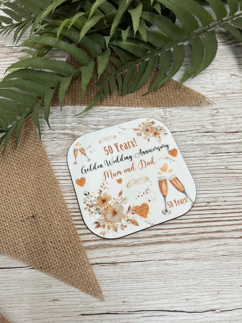 A 50th anniversary coaster with a floral design and champagne glasses, placed on a wooden surface with green leaves in the background.