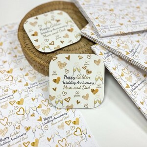 a collection of wedding anniversary coasters and greeting cards with a gold and white color scheme. The coasters and cards are adorned with gold hearts and text that reads "Happy Golden Wedding Anniversary, Mum and Dad" and "Happy Wedding Anniversary, Dad". The coasters and cards are arranged on a white surface, with some overlapping each other.