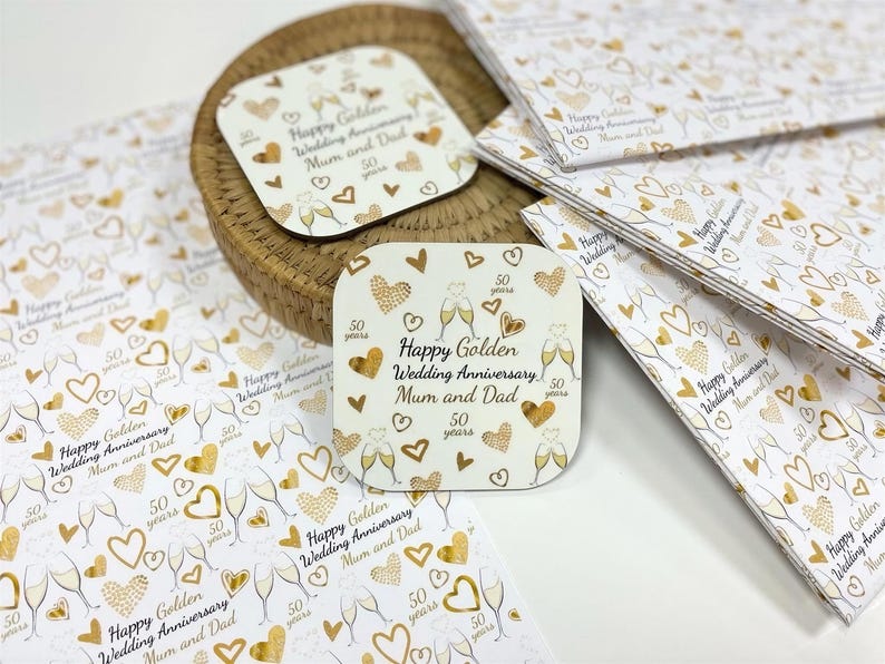 a collection of wedding anniversary coasters and greeting cards with a gold and white color scheme. The coasters and cards are adorned with gold hearts and text that reads "Happy Golden Wedding Anniversary, Mum and Dad" and "Happy Wedding Anniversary, Dad". The coasters and cards are arranged on a white surface, with some overlapping each other.
