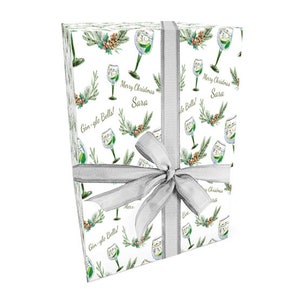 May include: Wrapped gift with white paper printed with gin glasses, pine branches, and the text "Gin-gle Bells!" and "Merry Christmas Sara." A silver ribbon and bow secure the present, ideal for Christmas.