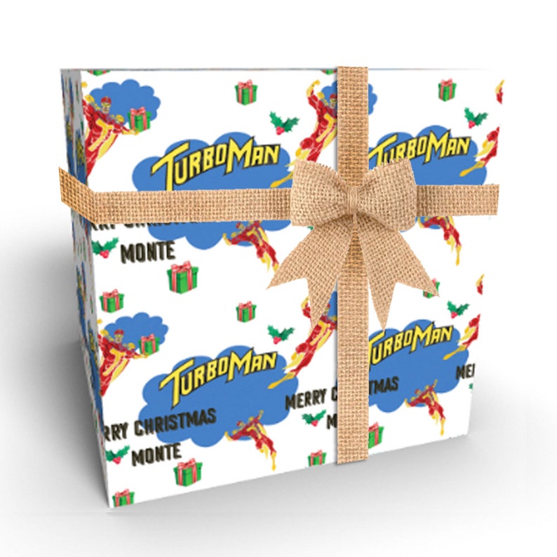 Puede incluir: Una caja de regalo envuelta con un patr&oacute;n repetido del personaje Turboman, cajas de regalo y acebo sobre un fondo blanco. La caja est&aacute; atada con una cinta y un lazo de arpillera. El texto en la caja dice "TurboMan" y "Merry Christmas Monte".