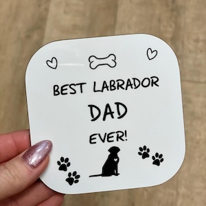 Może przedstawiać: Biały kwadratowy podkładka z czarnym tekstem i ilustracjami. Tekst brzmi "Best Labrador Dad Ever!" z kością, dwoma sercami i dwoma odciskami łap. Czarna sylwetka labradora retrievera znajduje się w środku.
