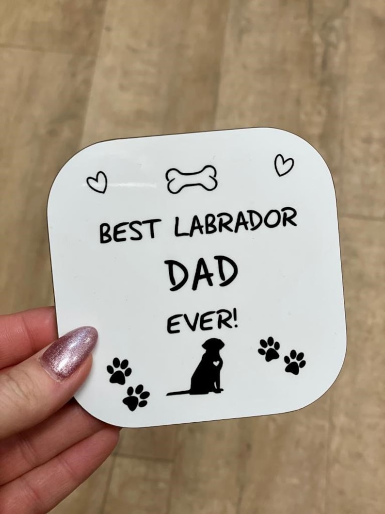 Może przedstawiać: Biały kwadratowy podkładka z czarnym tekstem i ilustracjami. Tekst brzmi "Best Labrador Dad Ever!" z kością, dwoma sercami i dwoma odciskami łap. Czarna sylwetka labradora retrievera znajduje się w środku.