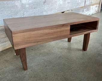 May include: A mid-century modern style coffee table made of walnut wood. The rectangular table has a recessed shelf and four tapered legs. The table is approximately 48 inches long, 16 inches wide, and 18 inches high.