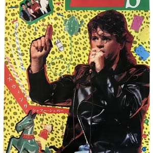 May include: A vintage INXS music poster featuring a person in a black leather jacket and blue jeans singing into a microphone. The yellow background has a black leopard print pattern, a small group photo, a green toy horse, and Japanese text.