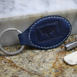 May include: A blue leather keychain with a stitched border and an embossed buffalo silhouette. The keychain is next to a leather working tool and a bone folder.