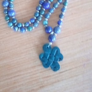 Puede incluir: Un collar de cuentas azules y verdes con un colgante verde oscuro. El collar tiene un diseño en capas con cuentas de diferentes tamaños. El colgante tiene una forma abstracta. Las cuentas son probablemente de piedra.